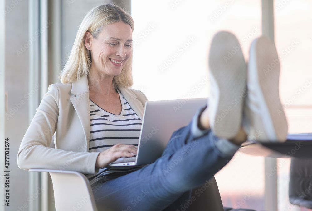 Smiling relaxed businesswoman using laptop in office with feet up