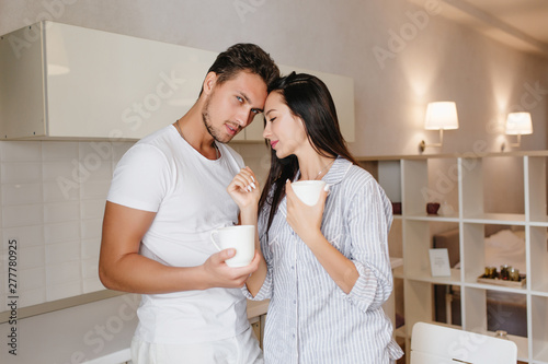 Sleepy black-haired girl standing in kitchen with boyfriend and drinking hot beverage. Married couple in cozy pajamas enjoys morning coffee. © Look!