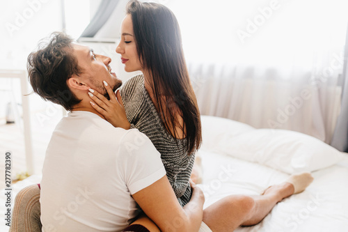 Enchanting girl with white manicure chilling in bed with husband and gently touching him. Indoor portrait of gorgeous lady in trendy knitted attire hugging with bearded man.