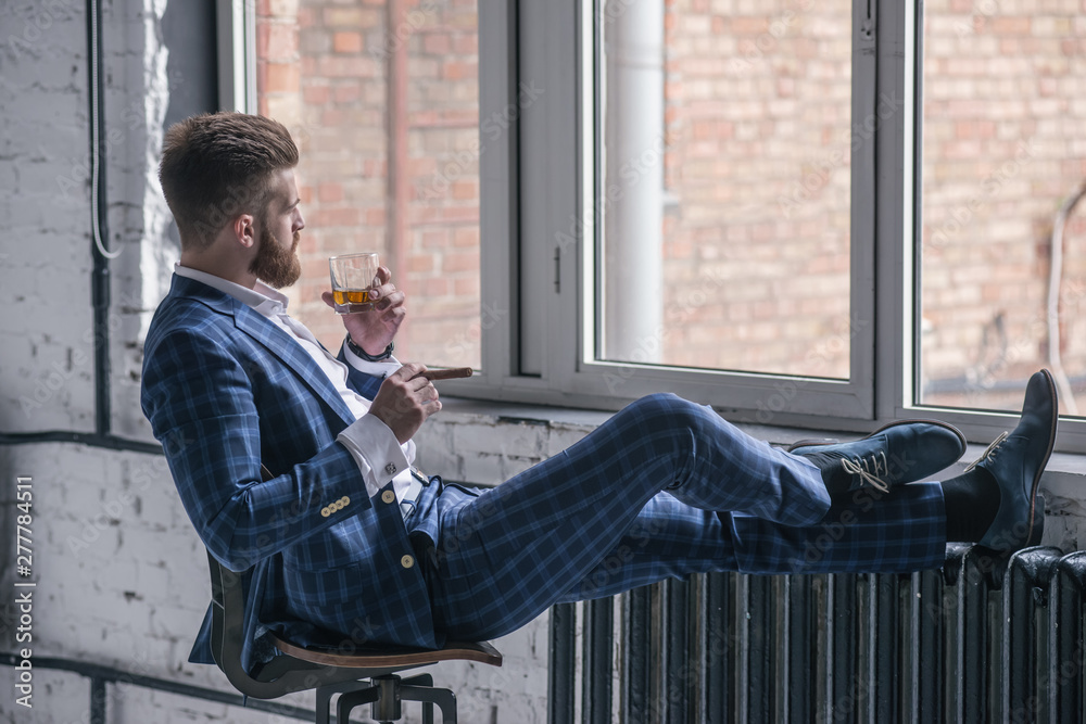 Handsome confident man is holding a cigar and a glass of whiskey and looking away while siting on the stool and looking to the window indoors. side view.