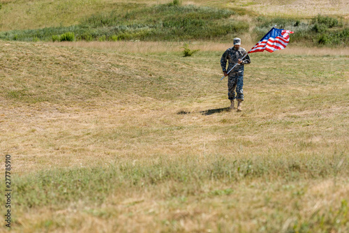selective focus of handsome soldier in uniform walking and holding american flag in summertime