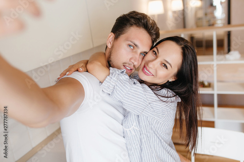 Ecstatic woman with brown eyes huggs with boyfriend while he making selfie. Indoor photo of caucasian brunette guy taking picture of himself with laughing european girl. photo