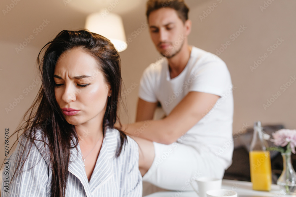 Upset girl with straight brown hair posing at home with sad man on background. Indoor portrait of unhappy married couple wear pajamas in morning.