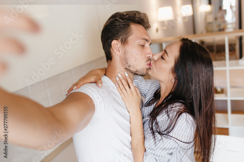 Blissful woman with elegant white manicure kissing boyfriend while he makes selfie. Handsome man with stylish haircut taking photo relaxing with wife at home.
