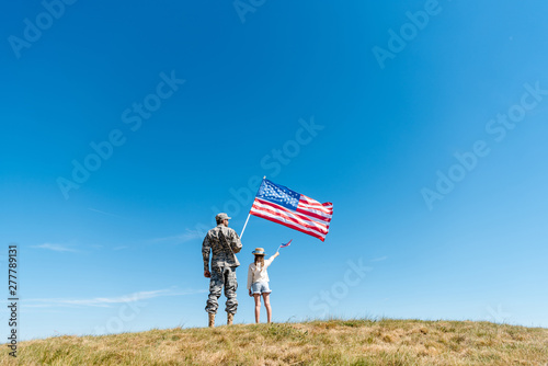 Back view of kid in straw hat and military father holding American flags