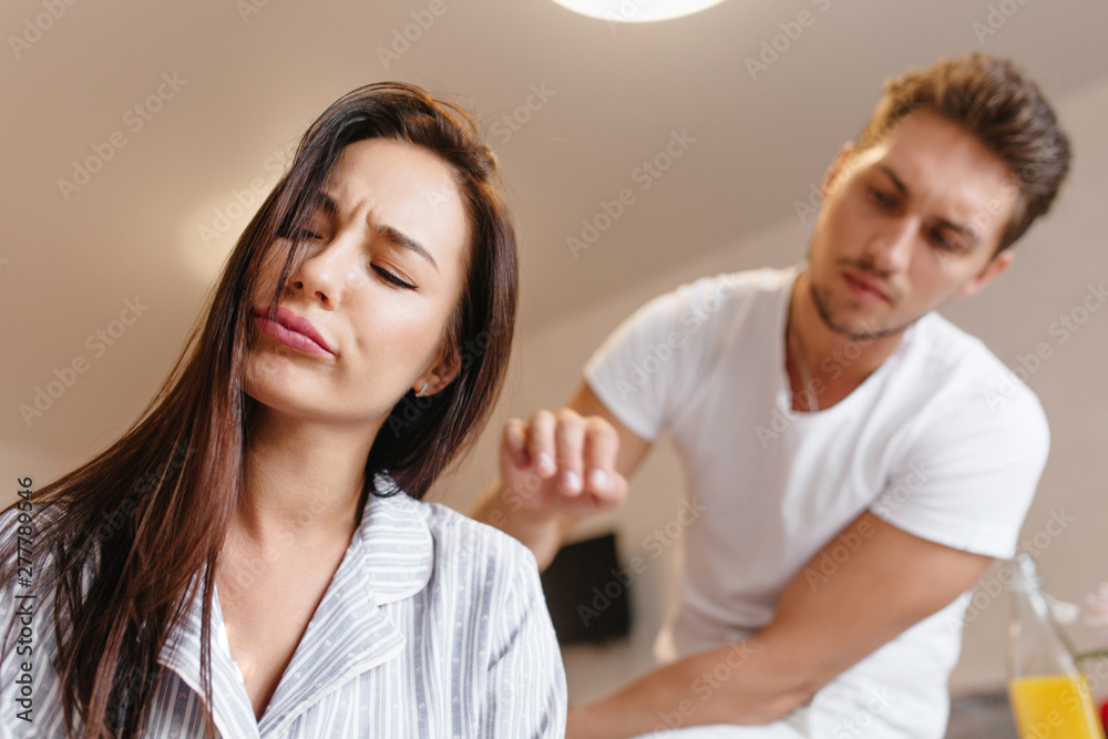 Upset female model in cute pajama posing with eyes closed while caucasian man telling something. Indoor portrait of frustrated brunette woman with her husband in white outfit on background. Upset female model in cute pajama posing with eyes closed while caucasian man telling something. Indoor portrait of frustrated brunette woman with her husband in white outfit on background.
