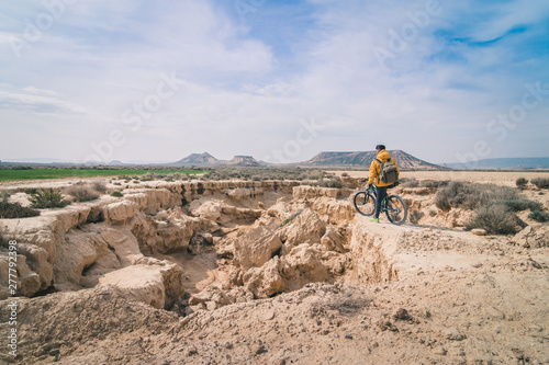 Side view of young man in yellow jacket and backpack with bicycle standing on stony hill stretchering high between stony hills in semi-desert Bardenas Reales Navarra Spain photo