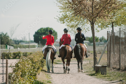 Back view of row of anonymous teen women riding horses in row strolling down roadway in sunlight photo