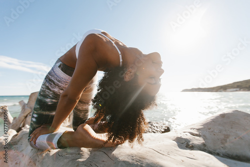 Side view of African American attractive young woman standing in yoga posture like bridge in open air on background of calm water in sunny day