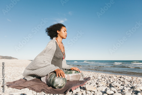 Side view of African American attractive young woman covered with grey plaid meditating in lotus yoga posture on sandy beach in bright day photo