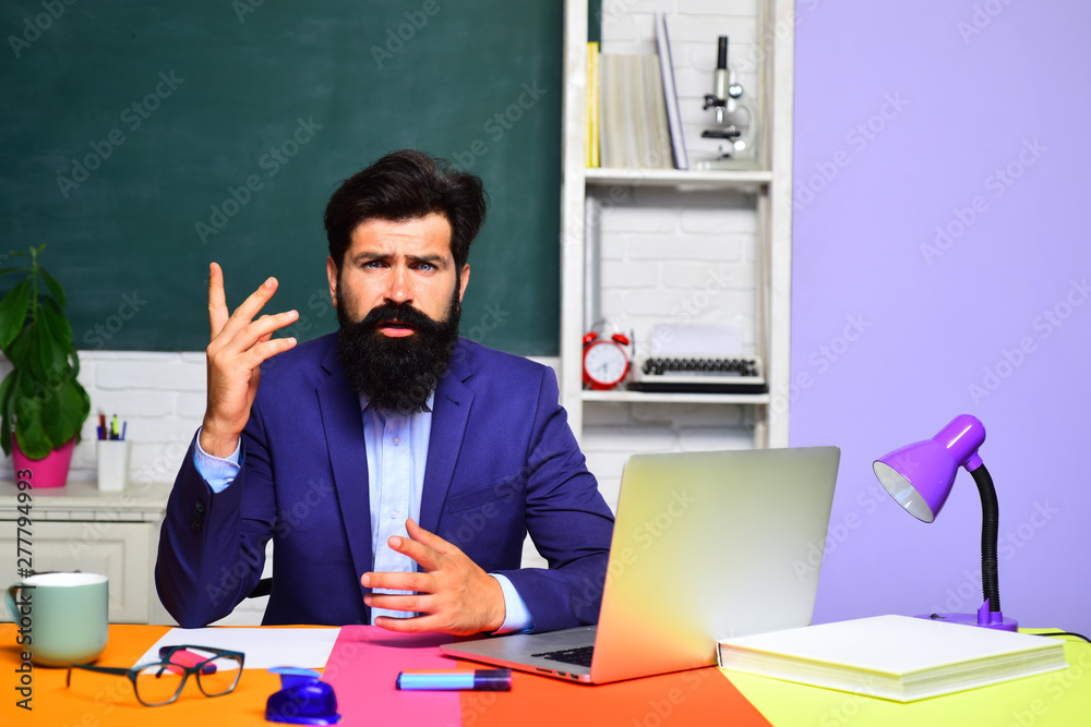 Teacher works with laptop in classroom. Hard work. Back to school. Online test. School, education, knowledge concept. Male teacher sits on desk. University student in glasses over green chalkboard. Teacher works with laptop in classroom. Hard work. Back to school. Online test. School, education, knowledge concept. Male teacher sits on desk. University student in glasses over green chalkboard.
