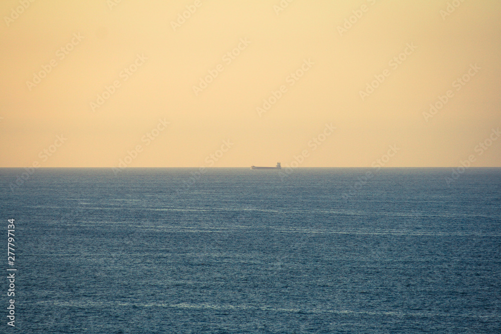 Landscape of an ocean with the horizon line centered on the image, a tanker sailing and an orange sky at sunset