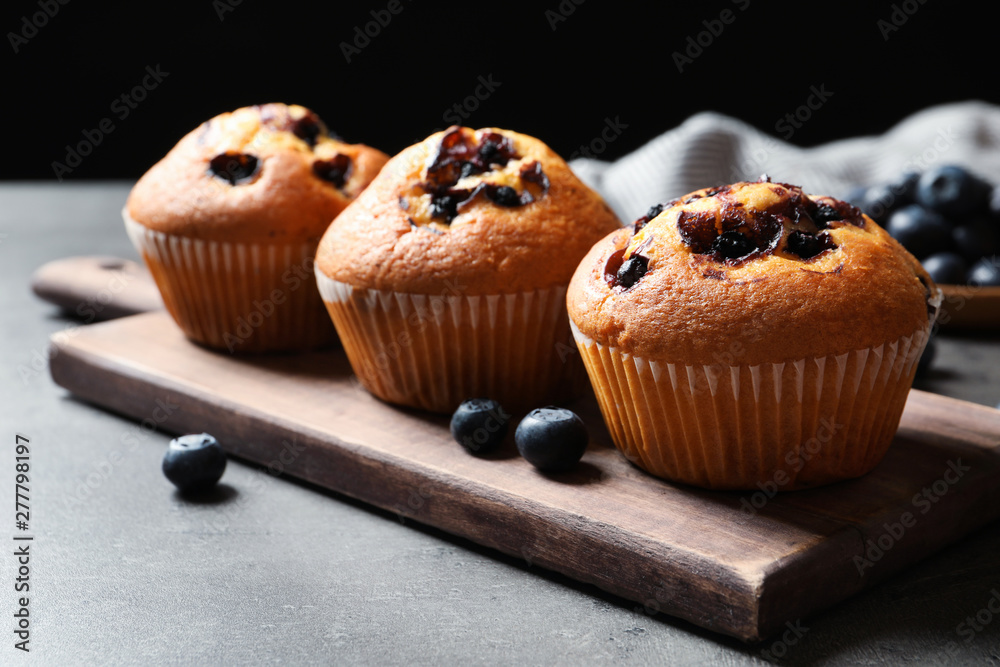 Wooden board with blueberry muffins on grey table against black background, closeup view Wooden board with blueberry muffins on grey table against black background, closeup view