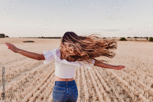 Summer Girl enjoying nature on yellow field. Beautiful young woman dancing Outdoors. Long hair in the wind. Happiness and lifestyle. Back view