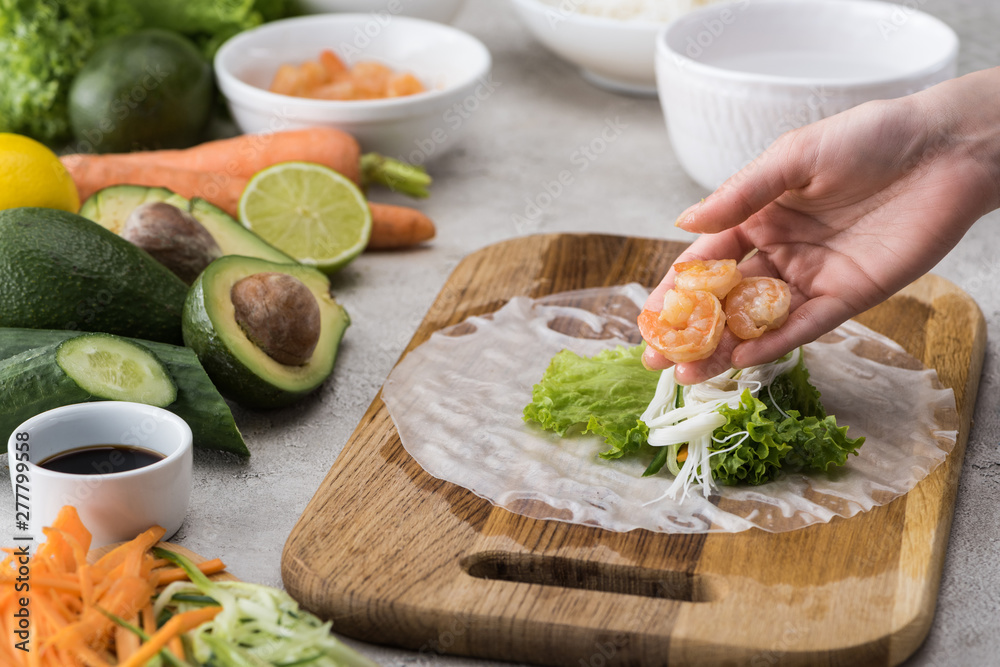 cropped view of woman putting shrimps on lettuce, noodles and rice paper on cutting board