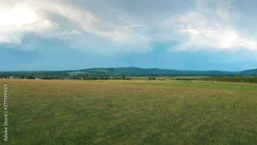 The panorama shot over the meadow. In the background is a village, forest and mountains. There are beautiful clouds in the sky. The panorama shot over the meadow. In the background is a village, forest and mountains. There are beautiful clouds in the sky.