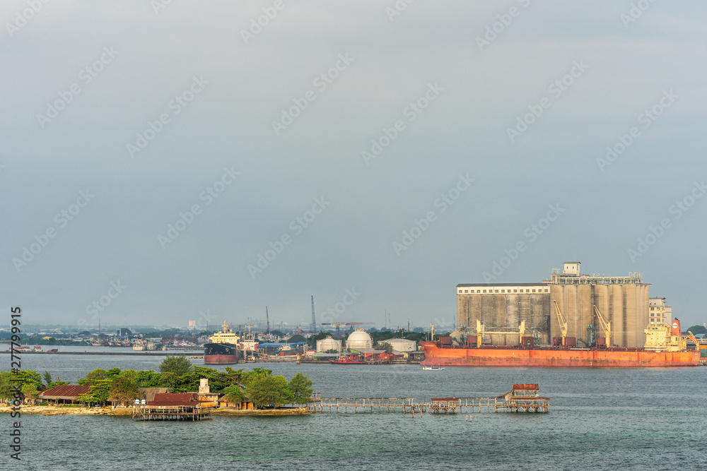 Makassar, Sulawesi, Indonesia - February 28, 2019: Evening shot of Pulau Khayangan island in front of the harbor. Brown concrete silos and large red Pan Begonia bulk carrier. City skyline.