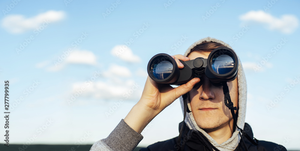 Obraz premium A man with modern binoculars against the sky and green hills. The concept of hunting, travel and outdoor recreation. Banner with copy space.