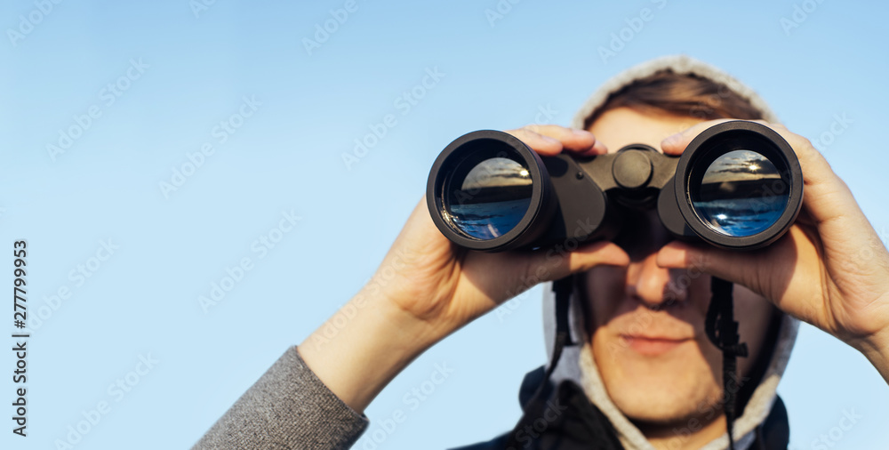 A man with modern binoculars against the sky and green hills. The concept of hunting, travel and outdoor recreation. Banner with copy space. A man with modern binoculars against the sky and green hills. The concept of hunting, travel and outdoor recreation. Banner with copy space.