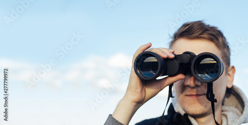 A man with modern binoculars against the sky and green hills. The concept of hunting, travel and outdoor recreation. Banner with  copy space.