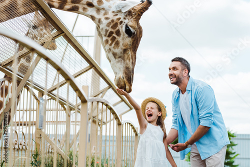Selective focus of cheerful man and kid with closed eyes feeding giraffe in zoo
