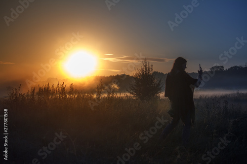 Silhouette of the traveler against fog landscape over a flower meadow  the first rays of dawn and dark silhouettes of trees against a sunrise  selective focus