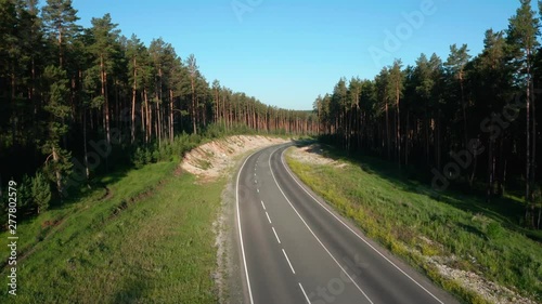 The panorama shot over the road. The highway passes through a dense forest. The forest is mostly coniferous. The sun illuminates the smooth road well.