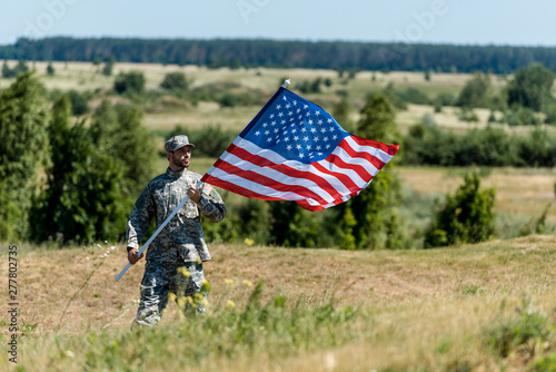 selective focus of handsome military man in uniform and cap holding american flag in summertime