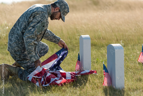 Konstfotografi soldier in uniform and cap holding american flag while sitting in graveyard