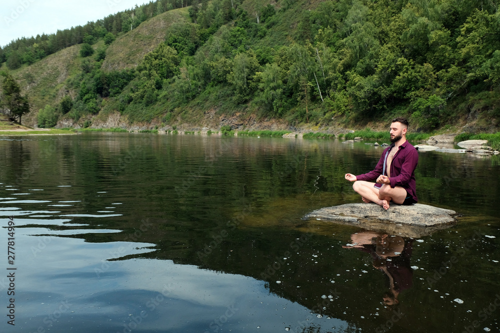 A guy with a kakvkazskoy appearance in a shirt with long sleeves sits on a stone in a river or lake. The concept of yoga in the forest, relaxation in the mountains, travel and lifestyle. Copy space.. A guy with a kakvkazskoy appearance in a shirt with long sleeves sits on a stone in a river or lake. The concept of yoga in the forest, relaxation in the mountains, travel and lifestyle. Copy space..
