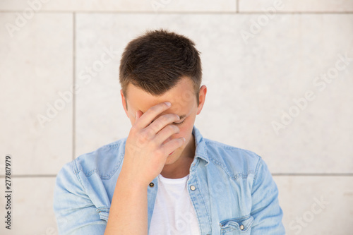 Tired young man touching forehead at wall outdoors. Handsome white guy wearing jeans shirt and keeping eyes closed with building wall in background. Stress or problem concept. Front view. © Mangostar