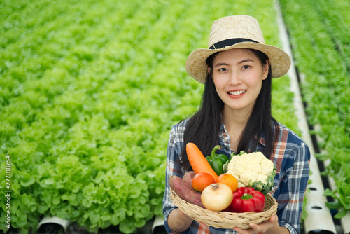 Young farmer girl holding various of vegetables in basket with smile. Organic vegetables ready to serve in salad dish for people who care about good health. Green vegetables farm in background.