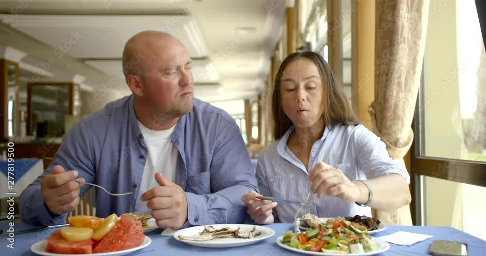 The husband bald and in glasses and the dark-haired middle-aged wife, sit in cafe, eat, treat each other, estimate food, laugh, communicate The husband bald and in glasses and the dark-haired middle-aged wife, sit in cafe, eat, treat each other, estimate food, laugh, communicate