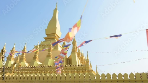 (Selective focus) Slow motion video of Laos flags waving in the foreground with the beautiful Pha That Luang in the background. Pha That Luang is a gold-covered large Buddhist stupa in Vientiane, Laos