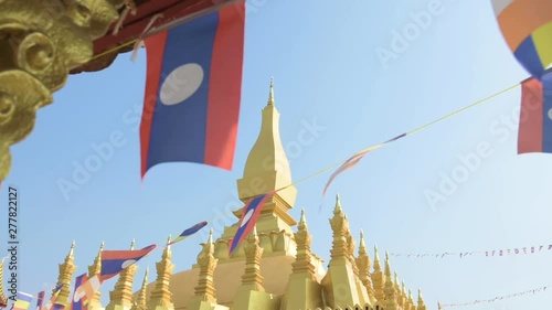 (Selective focus) Slow motion video of Laos flags waving in the foreground with the beautiful Pha That Luang in the background. Pha That Luang is a gold-covered large Buddhist stupa in Vientiane, Laos