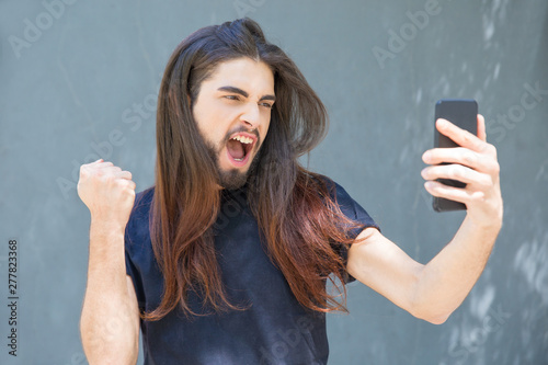 Bearded excited man with long hair taking selfie. Handsome young man with modern hairstyle making fist pump and photographing on smartphone. Success concept