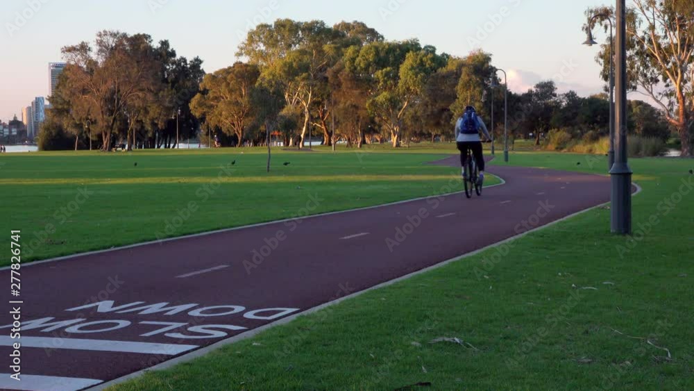 A bike rider zips along a winding track, late afternoon. A skateboard rider approaches from distance. Grass on either side, birds fossick in distance. Trees, people in background. River beyond. A bike rider zips along a winding track, late afternoon. A skateboard rider approaches from distance. Grass on either side, birds fossick in distance. Trees, people in background. River beyond.