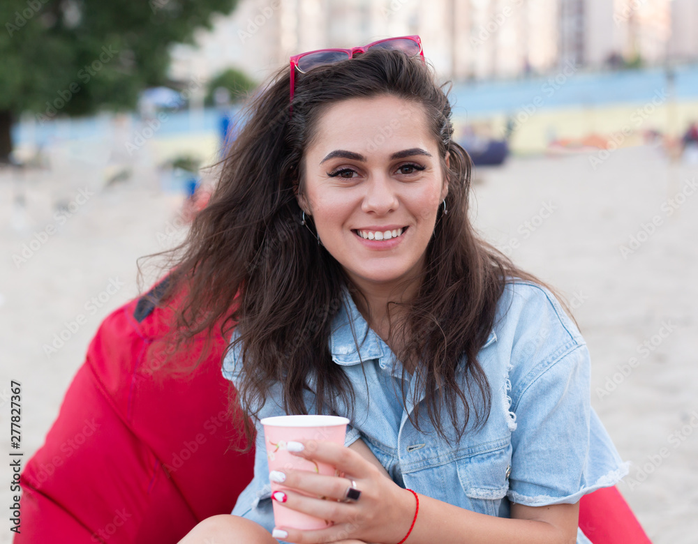 Young caucasian beautiful happy woman sitting on a red ottoman with cup of tea or coffee on the city beach in the summer. Youth concept