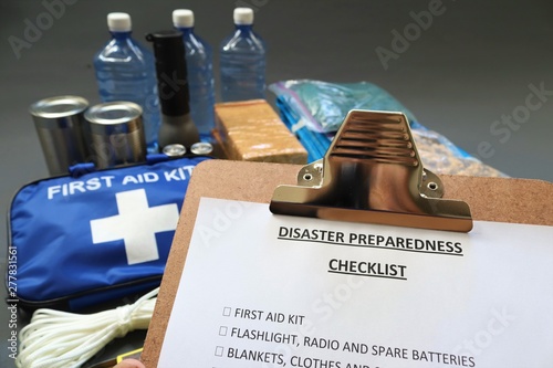 Disaster preparedness checklist on a clipboard with disaster relief items in the background.Such items would include a first aid kit,flashlight,tinned food,water,batteries and shelter.