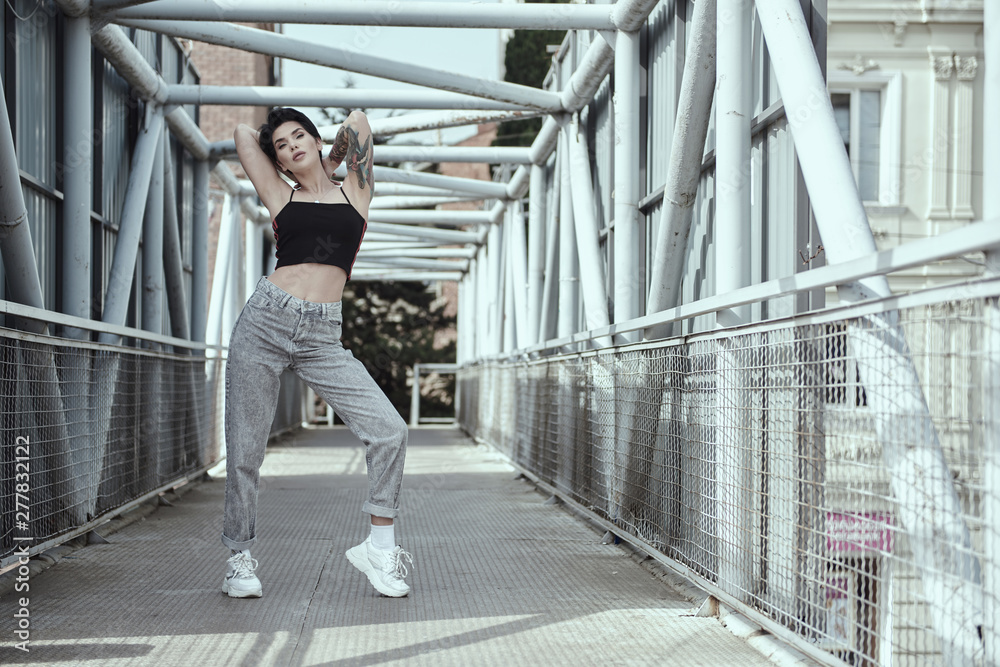 Raven haired female with tattoos is posing on the iron bridge in Tbilisi.  She wears top,