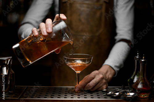 Male bartender pouring a brown alcoholic cocktail from the measuring cup to the glass