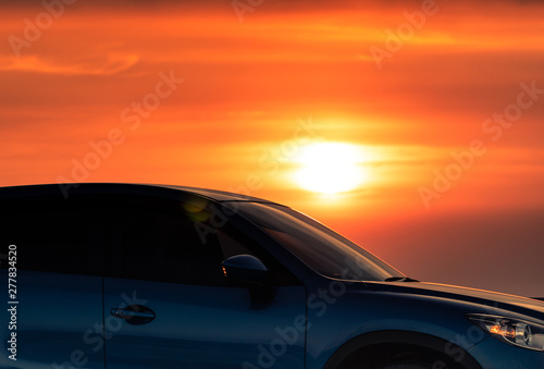 Side view of blue SUV car with sport and modern design parked on concrete road by sea beach at sunset. Electric car technology. Road trip travel. Automotive industry. Red and yellow sunset sky.  © Artinun