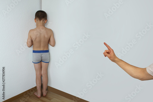 punished boy, a junior schoolboy, standing in a corner undressed, facing the wall, the hand of a parent faces a finger for misconduct, parenting concept