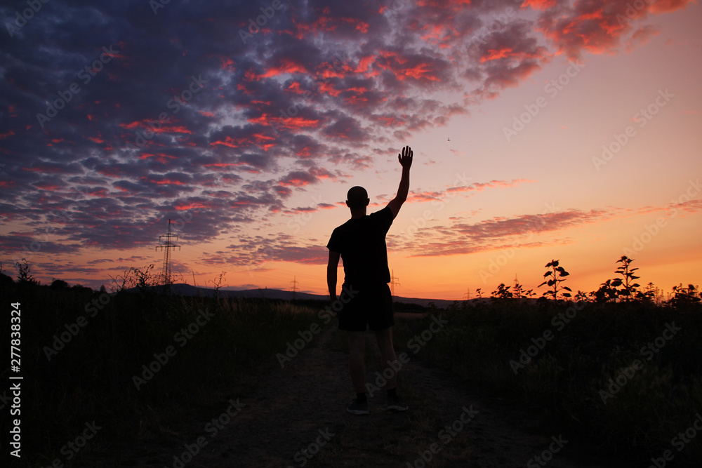 dark silhouette of a man in greeting raised one hand to the sky with a beautiful pink sunset with purple clouds
