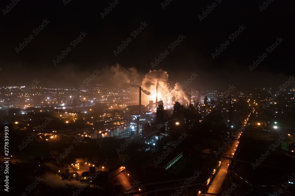 Aerial view of steel plant at night with smokestacks and fire blazing out of the pipe; industrial panoramic landmark with blast furnance of metallurgical production; concept of environmental pollution