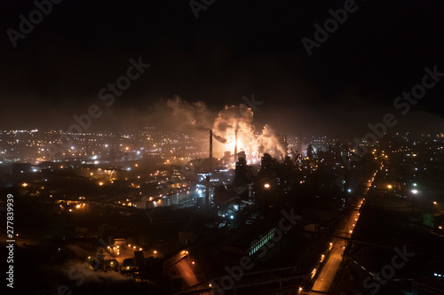 Aerial view of steel plant at night with smokestacks and fire blazing out of the pipe; industrial panoramic landmark with blast furnance of metallurgical production; concept of environmental pollution