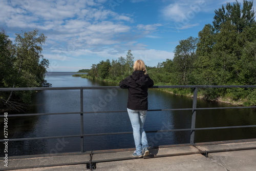 A girl stands on a bridge and looks into the distance. The river flows into the lake. Spring. Sunny day.