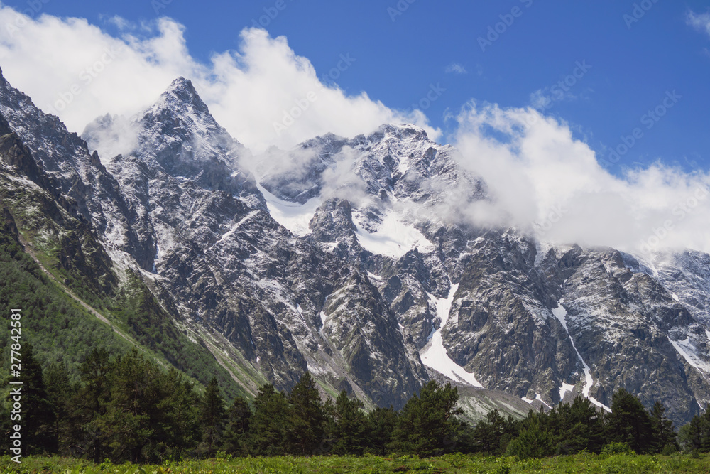 Snow-capped mountains in light clouds and warm green valley with small trees.