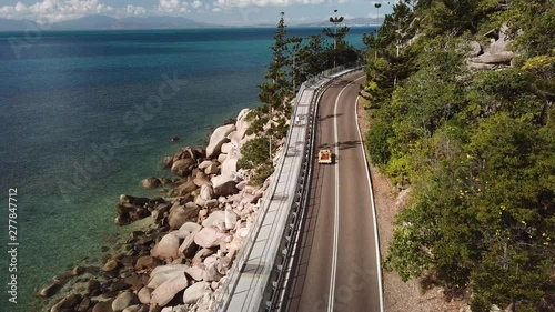 Aerial view of open top convertible classic beach buggy driving along coastal clifftop road next to crystal clear tropical water and lush green forest photo