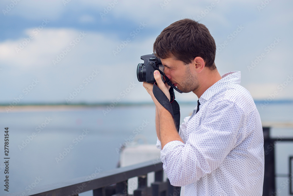 The young guy in a white shirt photographs the river from the embankment on the mirror camera